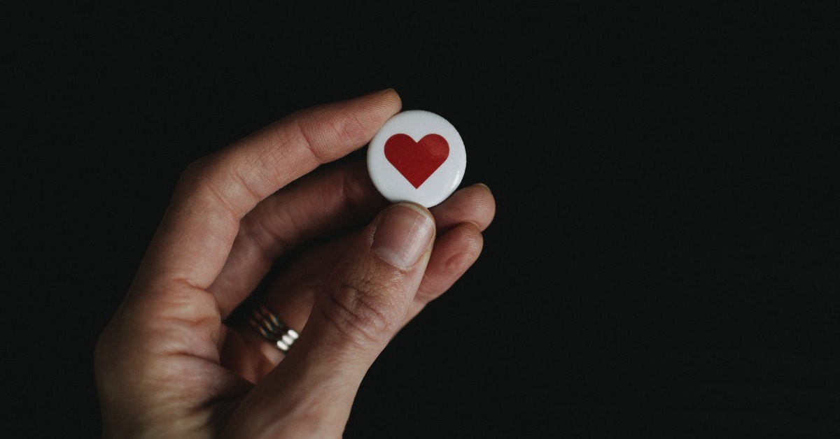 A person holding a white lapel pin with a red heart drawn on it 